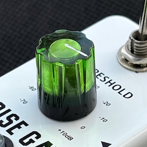 Può includere: Una manopola di controllo verde e scanalata con una linea indicatrice bianca è posizionata su una superficie bianca. La manopola è etichettata con numeri e la parola "THRESHOLD". È visibile anche il testo "NOISE GATE".