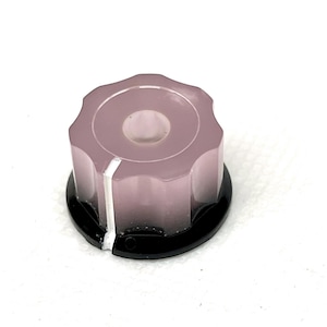 Op de afbeelding: Een doorschijnende, mauvekleurige knop met een witte indicatorlijn. De knop heeft een geribbelde, achthoekige vorm en staat op een zwarte, cirkelvormige basis. Deze bedieningsknop is waarschijnlijk voor een elektronisch apparaat.