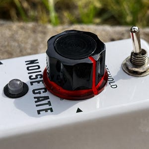 May include: A white guitar effects pedal labeled NOISE GATE with a large black control knob featuring a red translucent base. The pedal includes a silver metal toggle switch, a small circular indicator light, and a black input jack port.