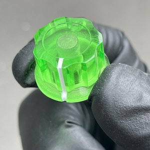 Könnte beinhalten: Ein neongrüner Bedienknopf mit einer weißen Markierungslinie. Der Knopf besteht aus transparentem Kunststoff und hat ein geriffeltes, kreisförmiges Design. Das Bild ist eine Nahaufnahme.