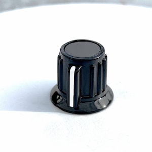 Könnte beinhalten: Schwarzer Bedienknopf mit weißer Markierungslinie. Der Knopf hat eine zylindrische Form mit vertikalen Rillen an den Seiten und einer flachen Oberseite. Der Sockel ist leicht ausgestellt, und der Knopf ist vor einem weißen Hintergrund platziert.