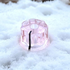 Op de afbeelding: Een heldere, roze, gefacetteerde knop met een zwarte indicatorlijn. De knop staat tegen een besneeuwde, witte achtergrond, waardoor een contrast ontstaat tussen het transparante object en het getextureerde oppervlak.