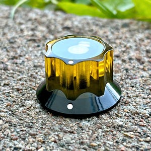 Peut inclure: Un bouton de commande transparent de couleur or avec une base noire. Le bouton a un dessus octogonal strié et un centre circulaire en retrait. Un petit point blanc est visible sur la base noire. Pour un appareil électronique.