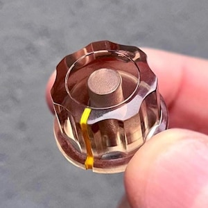 Pode incluir: Um botão de controlo de plástico castanho translúcido com uma borda estriada tipo engrenagem e um poste central cilíndrico. Uma única faixa amarela vertical está impressa na lateral do botão, estendendo-se da borda superior até à base.