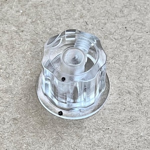 May include: A clear, transparent plastic knob featuring a multi-faceted, gear-like outer edge and a circular base. The knob has a central cylindrical post with internal threading and two small, distinct black dots marked on its surface.