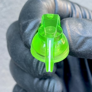 Può includere: Primo piano di un ugello di spruzzatore in plastica verde neon. L'ugello ha una base arrotondata e una parte superiore rettangolare con una striscia bianca al centro. L'ugello è tenuto da una mano guantata.
