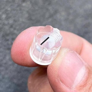 Pode incluir: Um pequeno objeto de plástico transparente com um exterior canelado e um design interior circular. Uma linha preta é visível através do centro do design circular. O objeto é segurado entre dois dedos.