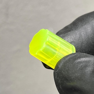 Puede incluir: Un objeto cilíndrico amarillo neón con un cuerpo estriado y una parte superior plana. El objeto está sostenido por una mano enguantada sobre un fondo neutro. El objeto parece estar hecho de un material translúcido.