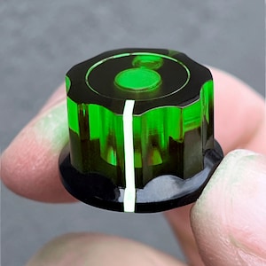 May include: A translucent green and black control knob with a white indicator line. The knob has a circular top with a recessed green center and a black base. The knob is held between fingers against a neutral background.