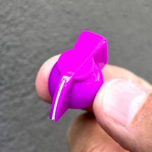 Könnte beinhalten: Ein leuchtend violetter Kunststoffknopf mit einer weißen Linie in der Mitte. Der Knopf ist für die Verwendung mit einer Gitarre oder einem anderen Musikinstrument gedacht.