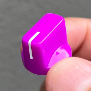 May include: A bright magenta plastic control knob featuring a smooth, glossy finish and a single vertical white indicator line on the top surface, designed for adjusting settings on electronic equipment or audio devices.