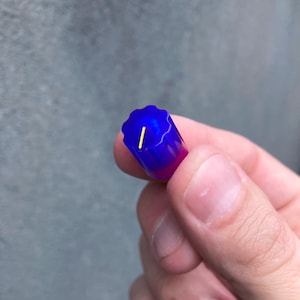 Puede incluir: Primer plano de una pequeña botella de plástico azul y morado con un pasador de metal dorado en su interior. La botella está siendo sostenida por una mano.