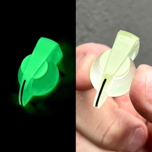 Może przedstawiać: Dwa świecące na zielono pokrętła sterowania z czarną linią wskaźnika. Jedno pokrętło na czarnym tle, drugie trzymane w dłoni na szarym tle. Pokrętła mają zaokrągloną podstawę i spiczastą górę.