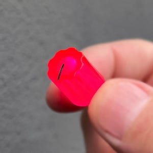 May include: A bright pink, cylindrical object with a scalloped edge and a black line. The object is held between fingers against a gray background. The object appears to be made of a translucent material.