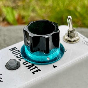 May include: A white guitar effects pedal featuring the text NOISE GATE in black lettering. The pedal includes a large black knob with a translucent teal base, a small silver toggle switch, and a circular indicator light on the top surface.