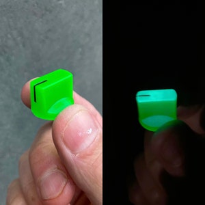 May include: A neon green, rectangular knob with a black line detail. The knob has a rounded base and appears to glow in the dark. The image shows the knob in two different lighting conditions.