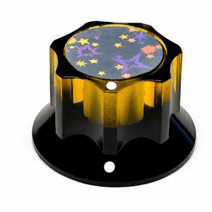 Peut inclure: Un bouton ambré translucide avec un bord cannelé à plusieurs côtés et un dessus circulaire plat. La surface supérieure présente un fond gris foncé décoré d'un motif de petites étoiles et de formes d'éclats jaunes, orange et violets.