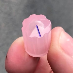 May include: A small, cylindrical, translucent light pink knob with a scalloped, fluted edge. The top surface features a recessed center with a single, thin, dark blue diagonal line indicator mark, providing a clear visual reference for orientation.
