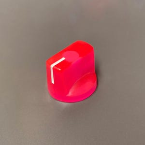 Puede incluir: Un mando de control rosa brillante con una línea indicadora blanca. El mando tiene una forma redondeada y probablemente se utiliza para ajustar la configuración de un dispositivo electrónico. El mando está hecho de un material sólido.