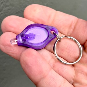 Puede incluir: Un peque&ntilde;o llavero con linterna de color morado, con una lente transparente y un llavero plateado. La linterna tiene forma de l&aacute;grima y tiene una peque&ntilde;a luz LED en el lateral.