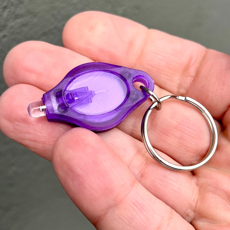 Puede incluir: Un peque&ntilde;o llavero con linterna de color morado, con una lente transparente y un llavero plateado. La linterna tiene forma de l&aacute;grima y tiene una peque&ntilde;a luz LED en el lateral.