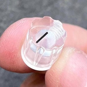 May include: A small, cylindrical, clear plastic cap featuring a scalloped top edge and a flat, circular white interior base. A single, thin, straight black line is printed across the center of the white circular surface of this component.