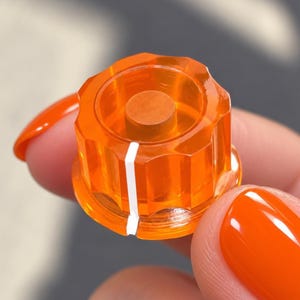 Puede incluir: Un mando de control circular de color naranja translúcido con un borde exterior estriado y una única línea indicadora blanca vertical. El centro del mando contiene una pieza circular naranja sólida, ofreciendo una vista clara de la estructura.