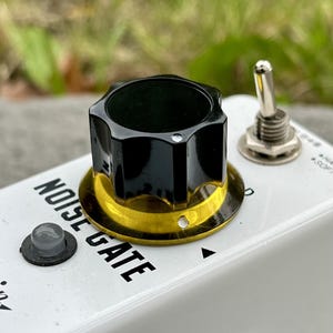Puede incluir: Un pedal de efectos de guitarra blanco con la etiqueta NOISE GATE, que presenta una perilla negra grande con una base dorada, una pequeña luz indicadora gris, un interruptor de palanca de metal plateado y un puerto jack circular en el chasis.