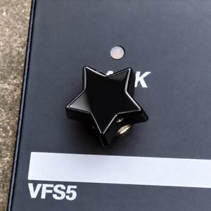 Könnte beinhalten: Ein glänzender, sternförmiger schwarzer Drehknopf auf einem mattschwarzen Bedienfeld. Das Panel zeigt eine kleine kreisförmige Kontrollleuchte, den Buchstaben K und den Text VFS5, der unter einem weißen horizontalen Streifen aufgedruckt ist.