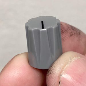 May include: A gray plastic control knob with a fluted, cylindrical body and a scalloped top edge. A single thin black vertical line is printed on the top surface to indicate position. The knob features a tapered design with vertical ridges.