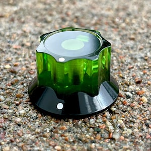 May include: A green and black control knob with a clear, faceted top. The knob has a circular design with a white dot on the black base. The top is a vibrant green colour, and the knob appears to be made of plastic.