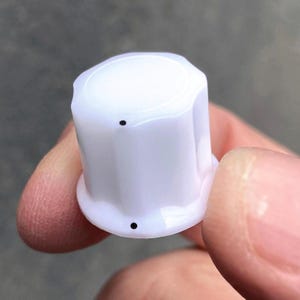 May include: A white cylindrical control knob with a fluted exterior surface. The knob features a flat top and a slightly wider base, with two small, distinct black circular markings positioned vertically on the side to indicate orientation.