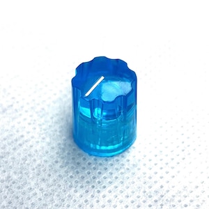 Könnte beinhalten: Ein durchscheinender blauer Knopf mit geriffelter, gezackter Oberseite und einer weißen Linie. Der zylindrische Knopf besteht aus einem transparenten Material, und die weiße Linie ist ein visueller Indikator. Der Knopf ist wahrscheinlich für ein elektronisches Gerät.