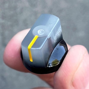 May include: A translucent gray control knob with a solid black circular base. The top surface features a bright yellow vertical indicator line and a circular indentation in the center. The knob has a smooth, rounded rectangular shape.