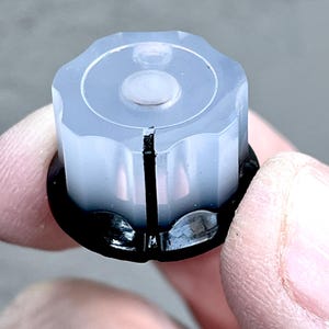 May include: A white plastic knob with a black centre and a small white button in the centre. The knob has a textured surface and is designed for use with a rotary control.