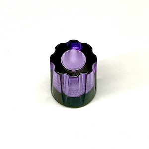 Op de afbeelding: Een kleine, cilindrische kandelaar met een donkerpaarse basis en een lichtere paarse bovenkant. De bovenkant heeft een geschulpte rand en het midden is een cirkelvormige opening voor een kaars. De kandelaar is van glas.