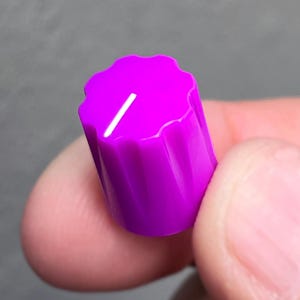 May include: A vibrant magenta control knob featuring a scalloped, fluted edge design and a single white indicator line on the top surface. The cylindrical component has a textured grip for manual adjustment on electronic equipment or audio gear.