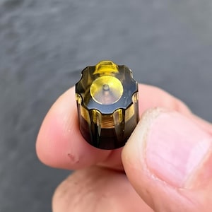 Op de afbeelding: Close-up van een klein, cilindrisch object met een bloemvormige bovenkant. Het object is voornamelijk zwart met gouden accenten en een transparant geel centrum. Het object wordt tussen twee vingers gehouden.