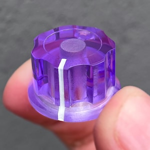 May include: A translucent purple plastic control knob with a cylindrical center and a fluted outer edge. A single vertical white line is printed on the side of the knob to indicate position, and the top surface features a circular recessed detail.