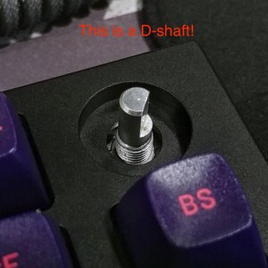 Puede incluir: Primer plano de un interruptor de teclado mec&aacute;nico plateado con un v&aacute;stago en forma de D. El interruptor est&aacute; montado en una placa de teclado negra y rodeado de teclas moradas. La tecla "BS" es visible en primer plano.