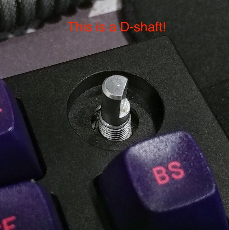 Puede incluir: Primer plano de un interruptor de teclado mec&aacute;nico plateado con un v&aacute;stago en forma de D. El interruptor est&aacute; montado en una placa de teclado negra y rodeado de teclas moradas. La tecla "BS" es visible en primer plano.