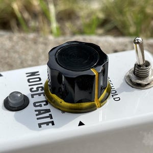 May include: A white guitar effects pedal labeled NOISE GATE with a black control knob featuring a yellow indicator line, a metallic toggle switch, and a small circular indicator light. The text THRESHOLD is partially visible on the white surface.