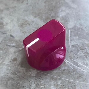 May include: A glossy magenta control knob featuring a smooth, rounded top surface with a single white indicator line extending from the center toward the edge. The knob has a tapered cylindrical body with a polished finish and a flat top.