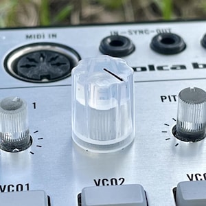 May include: A clear, fluted plastic control knob with a black indicator line sits on a silver synthesizer panel. The panel features black text labels including MIDI IN, SYNC, and VCO2, along with additional smaller knobs and grey rectangular buttons.