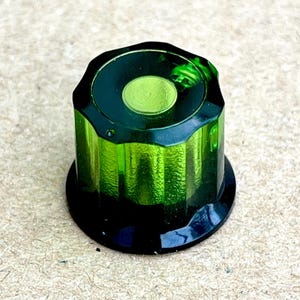 Puede incluir: Un mando de control verde esmeralda translúcido con un cuerpo cilíndrico estriado y una parte superior circular lisa. El mando presenta una base circular negra sólida y un pequeño tornillo metálico interno visible a través del plástico.