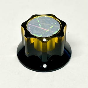 Puede incluir: Un mando de control de color ámbar translúcido con un borde estriado de varios lados y una superficie superior plana. La parte superior presenta una incrustación oscura con acentos lineales iridiscentes y multicolores. El mando se asienta sobre una base negra.