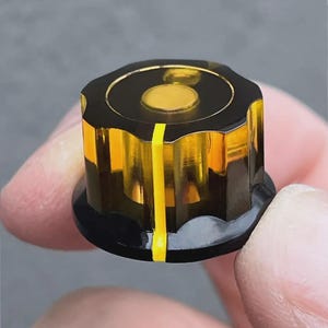 Könnte beinhalten: Ein schwarzer und bernsteinfarbener Knopf mit einem gelben Streifen in der Mitte. Der Knopf hat eine erhabene, kreisförmige Mitte.