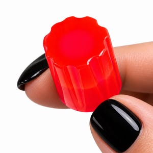 May include: A small, translucent, bright red cylindrical cap with a fluted, scalloped edge design. The object features a smooth, glossy finish and a distinct ridged texture along its vertical sides, creating a uniform, repeating pattern.