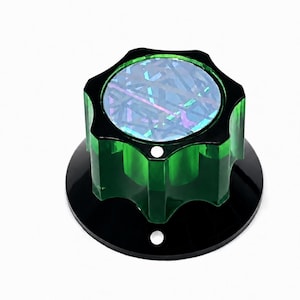 Puede incluir: Un pomo verde translúcido con borde festoneado y parte superior plana que presenta un patrón geométrico holográfico multicolor. El pomo está unido a una placa base circular negra que tiene dos pequeños orificios circulares espaciados.