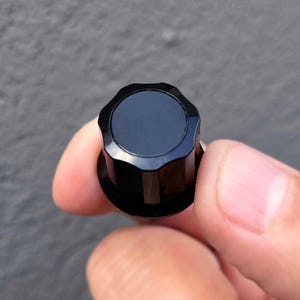 以下が含まれることがあります： 円形のトップと波状の溝があるエッジを備えた黒いコントロールノブ。ノブは滑らかで平らな中央表面と光沢のある仕上げが特徴で、電子機器やオーディオデバイスの調整用に設計されています。円筒形のベースが付いています。